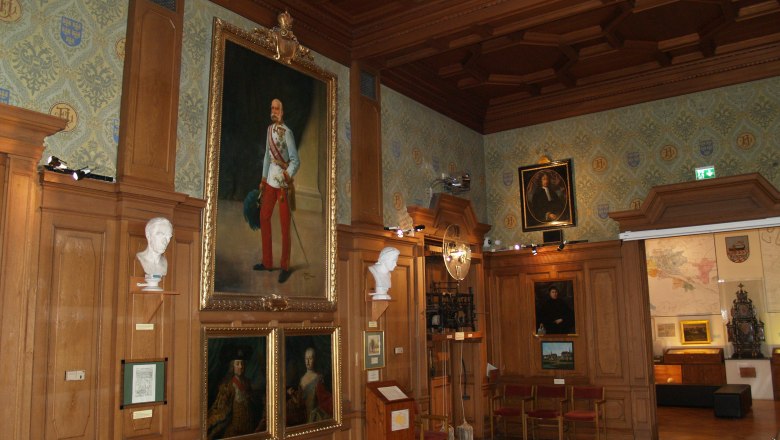 A historic hall with wood paneling, paintings and busts on the walls.