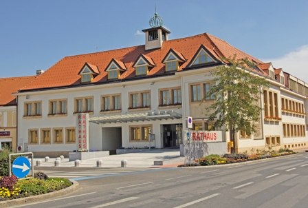 Rathaus Traiskirchen mit rotem Dach und mehreren Fenstern, umgeben von einer Straße und Blumenbeeten.