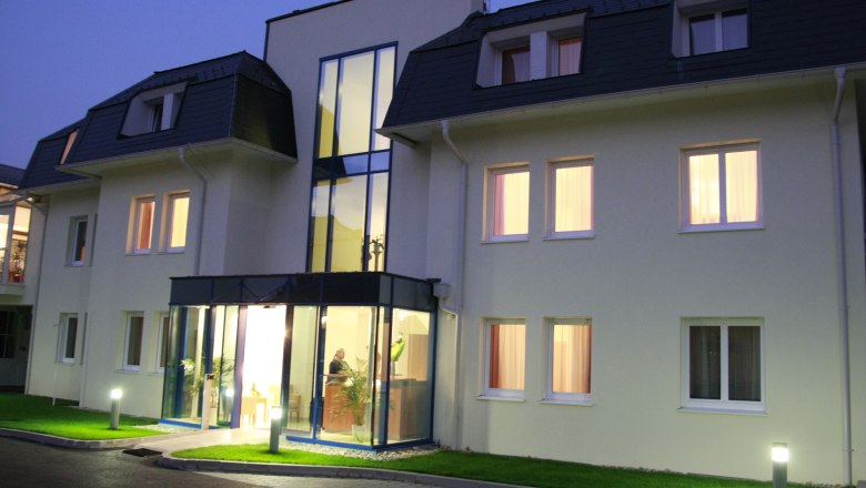 Eingangsbereich eines modernen Hotels bei D&auml;mmerung mit beleuchteten Fenstern und Glasfassade.