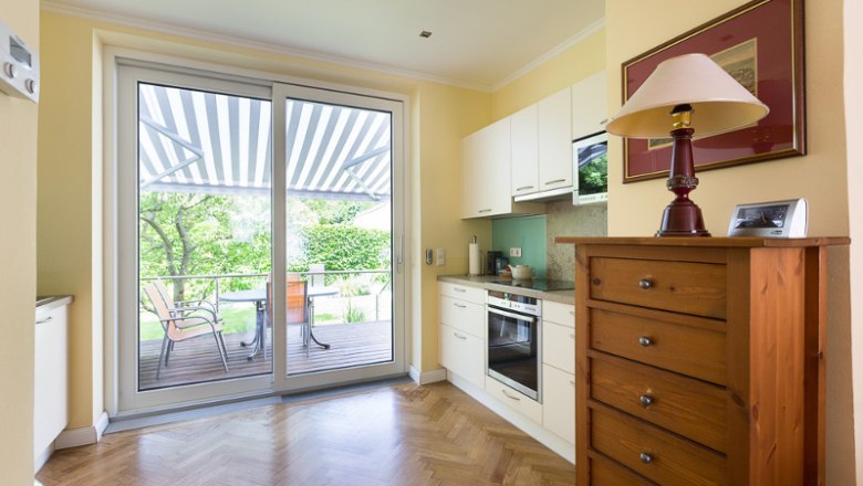 Helle K&uuml;che mit Parkettboden, Schrank, Herd und gro&szlig;er Glast&uuml;r zur Terrasse mit Markise.