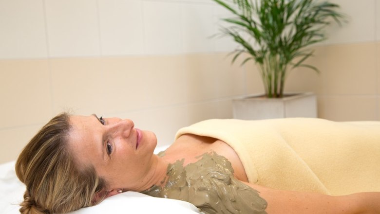 Woman relaxing with a sulphur mud pack on her shoulder, wrapped in a blanket, in the wellness area with a plant in the background.