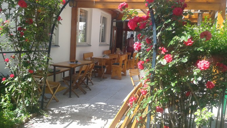 Ein Rosenbogen mit roten Rosen f&uuml;hrt zu einem Au&szlig;enbereich mit Holztischen und -st&uuml;hlen.