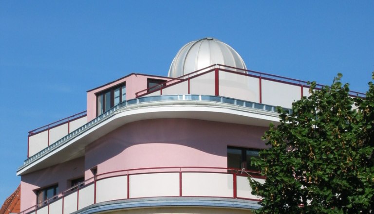 Ein modernes Gebäude mit einer Kuppel auf dem Dach, umgeben von Bäumen, unter einem klaren blauen Himmel.