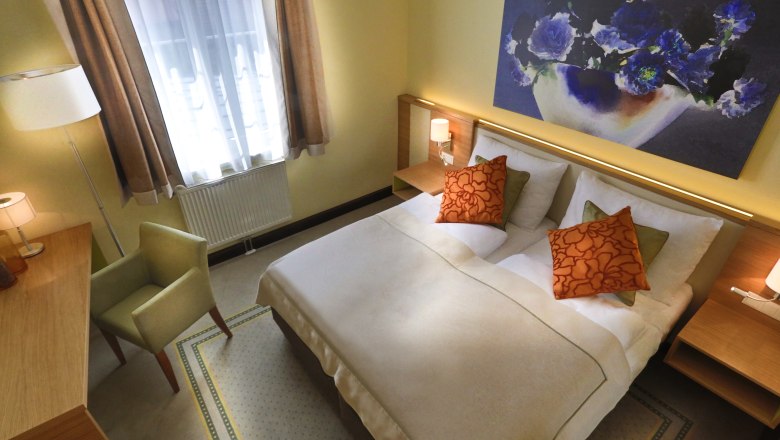 Cozy hotel room with a double bed, two orange pillows, a desk, an armchair and a large flower picture on the wall.