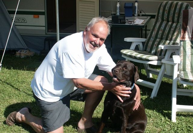 A man kneels next to a brown dog on a meadow in front of a caravan.
