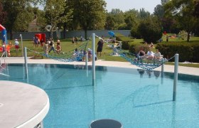 Ein Freibad mit Hängematten über dem Wasser und Menschen im Hintergrund auf der Liegewiese.