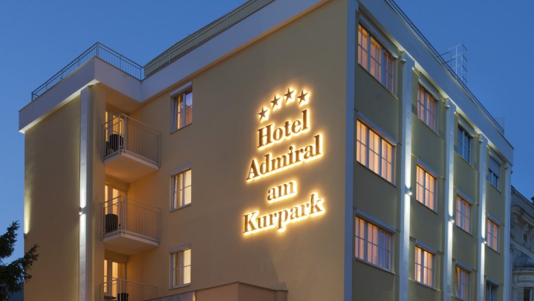 Au&szlig;enansicht eines Hotels bei D&auml;mmerung mit beleuchtetem Schriftzug "Hotel Admiral am Kurpark".