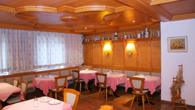 Gem&uuml;tlicher Fr&uuml;hst&uuml;cksraum mit Holzm&ouml;beln, rosa Tischdecken und dekorativen Kr&uuml;gen auf einem Regal.