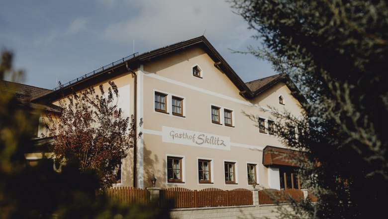 Ein traditionelles Gasthaus mit der Aufschrift 'Gasthof Skilitz', umgeben von B&auml;umen und einem Holzzaun.