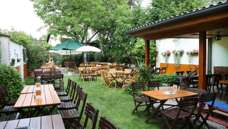 Ein gemütlicher Biergarten mit Holztischen und -stühlen, umgeben von grünen Bäumen und Pflanzen.