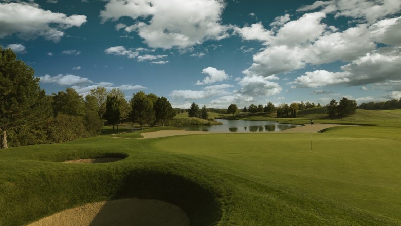 Golfplatz mit Sandbunker, Grün und Teich unter blauem Himmel mit Wolken.