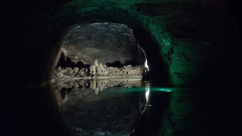 Dunkle H&ouml;hle mit Wasser und Felsformationen, beleuchtet von gr&uuml;nlichem Licht.