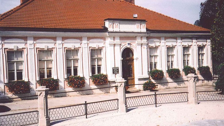 Historic building with red tiled roof and white window frames, surrounded by flowers.