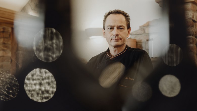 Ein Mann in schwarzer Kochkleidung steht in einem rustikalen Raum, umgeben von unscharfen Lichteffekten.