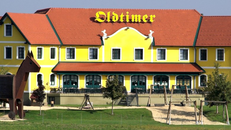 Gelbes Gebäude mit rotem Dach und der Aufschrift 'Oldtimer', daneben ein Spielplatz mit einer großen Holzfigur.