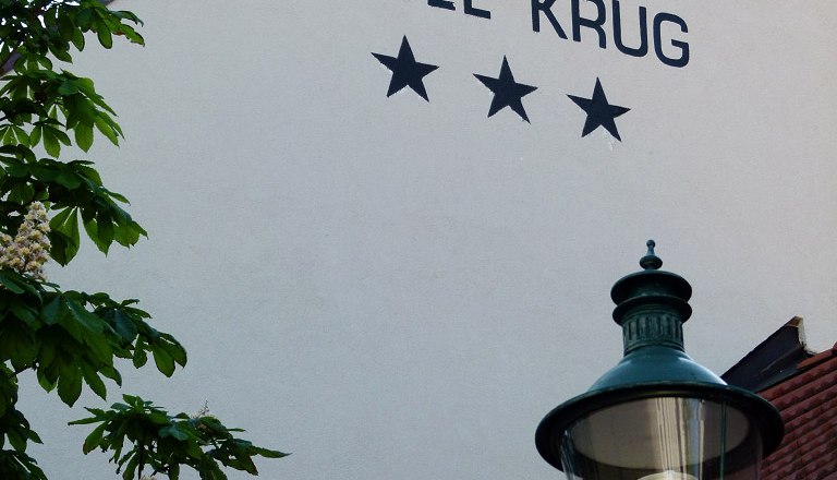Fassade des Hotel Krug mit drei Sternen, Baum und Straßenlaterne.